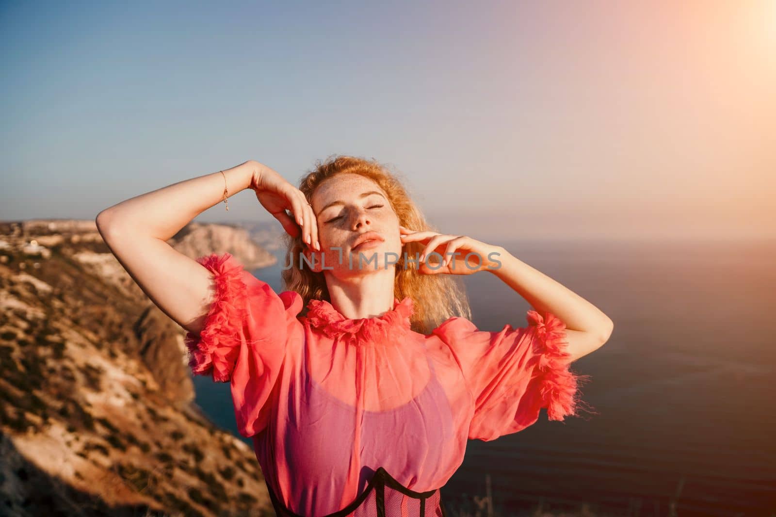 Close up shot of beautiful young caucasian woman with curly blond hair and freckles looking at camera and smiling. Cute woman portrait in a pink long dress posing on a volcanic rock high above the sea
