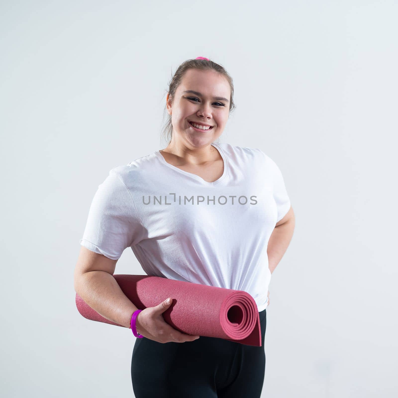 Young fat caucasian woman holding a sport mat. Charming plus size model in sportswear stands on a white background by mrwed54