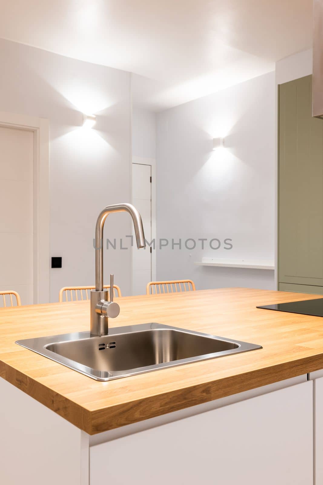 Close-up of sink in wooden countertop in kitchen table. Mixer and sink in chromed metal. The kitchen is lit with bright designer wall lights. Kitchen area at the entrance to studio apartment