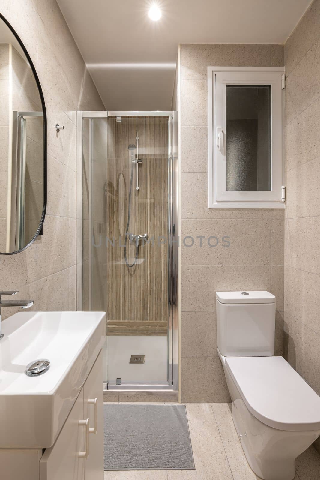 Compact bathroom with bright lighting from ceiling. Beige granite tiles on walls. White toilet bowl and sink on dressing table, shower area with transparent glass partition. Oval mirror on wall