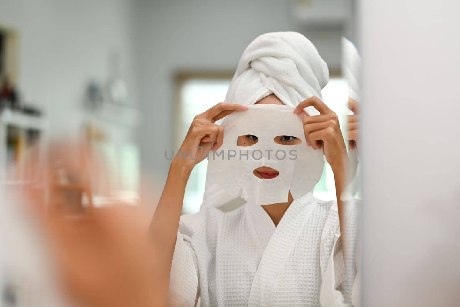 Mirror reflection of woman wearing bath towel on head applying in sheet face mask. Beauty treatment concept.