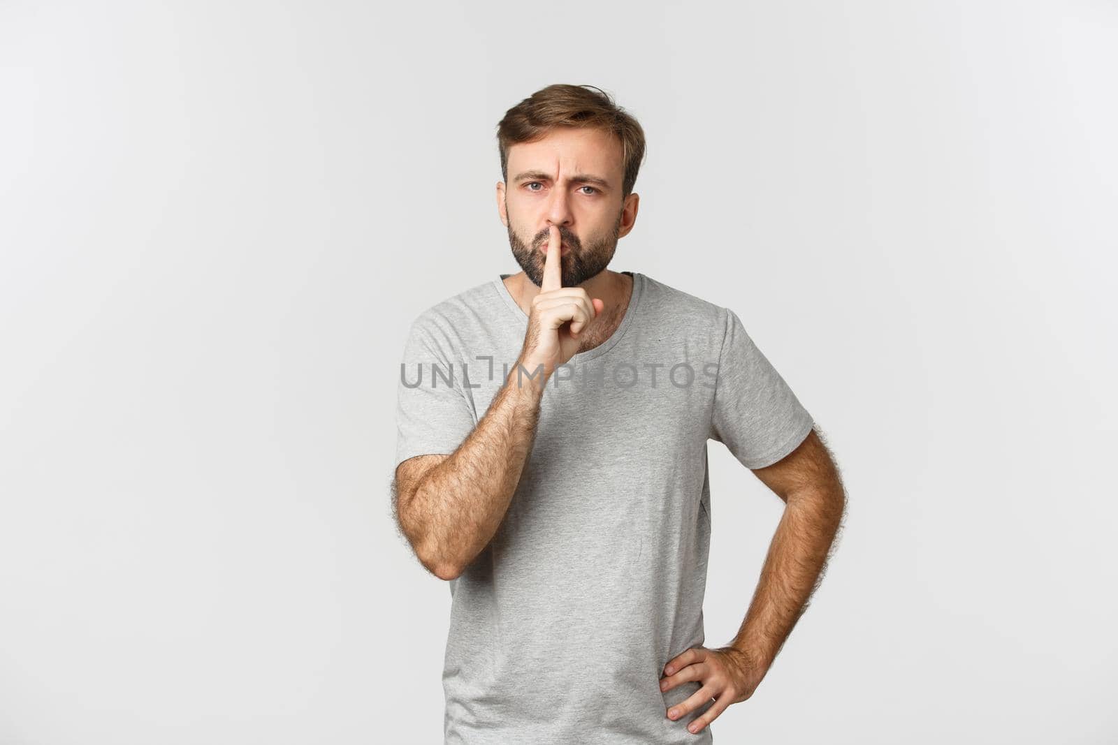 Angry bearded man in grey t-shirt, hushing and frowning, scolding person for talking too loud, standing over white background by Benzoix