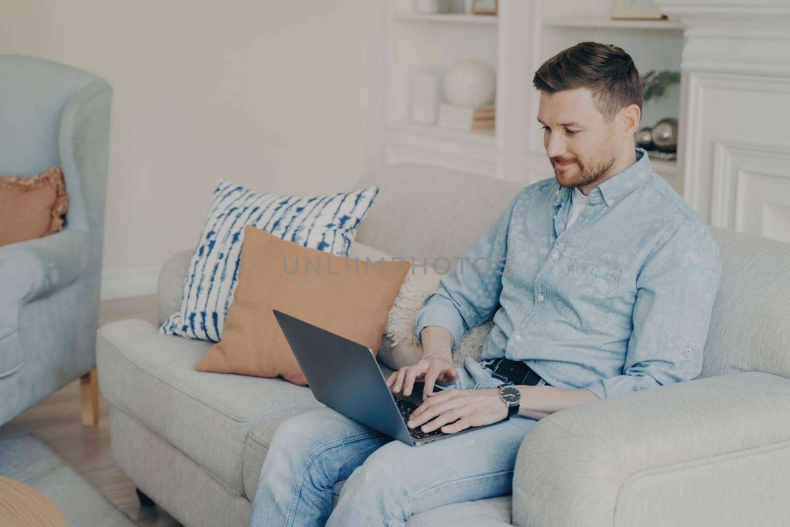 Relaxed casually dressed young businessman working from home while sitting on comfortable couch in light living room, typing on laptop and using special business software. Freelance concept