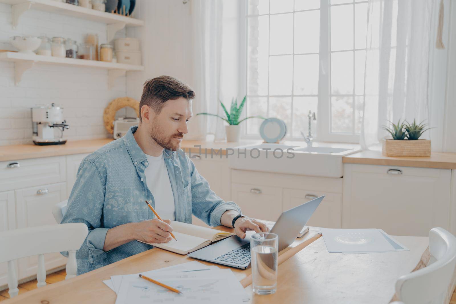 Serious man working on laptop online, sitting at kitchen table and looking at computer screen, focused male writing notes while studying online or searching information for his reports