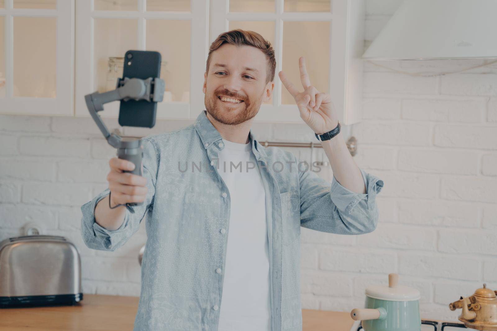 Handsome smiling young brunette man with stubble making selfie or streaming live on smartphone with gimbal stabilizer and gesturing peace sign in kitchen background at home, wears casual clothes