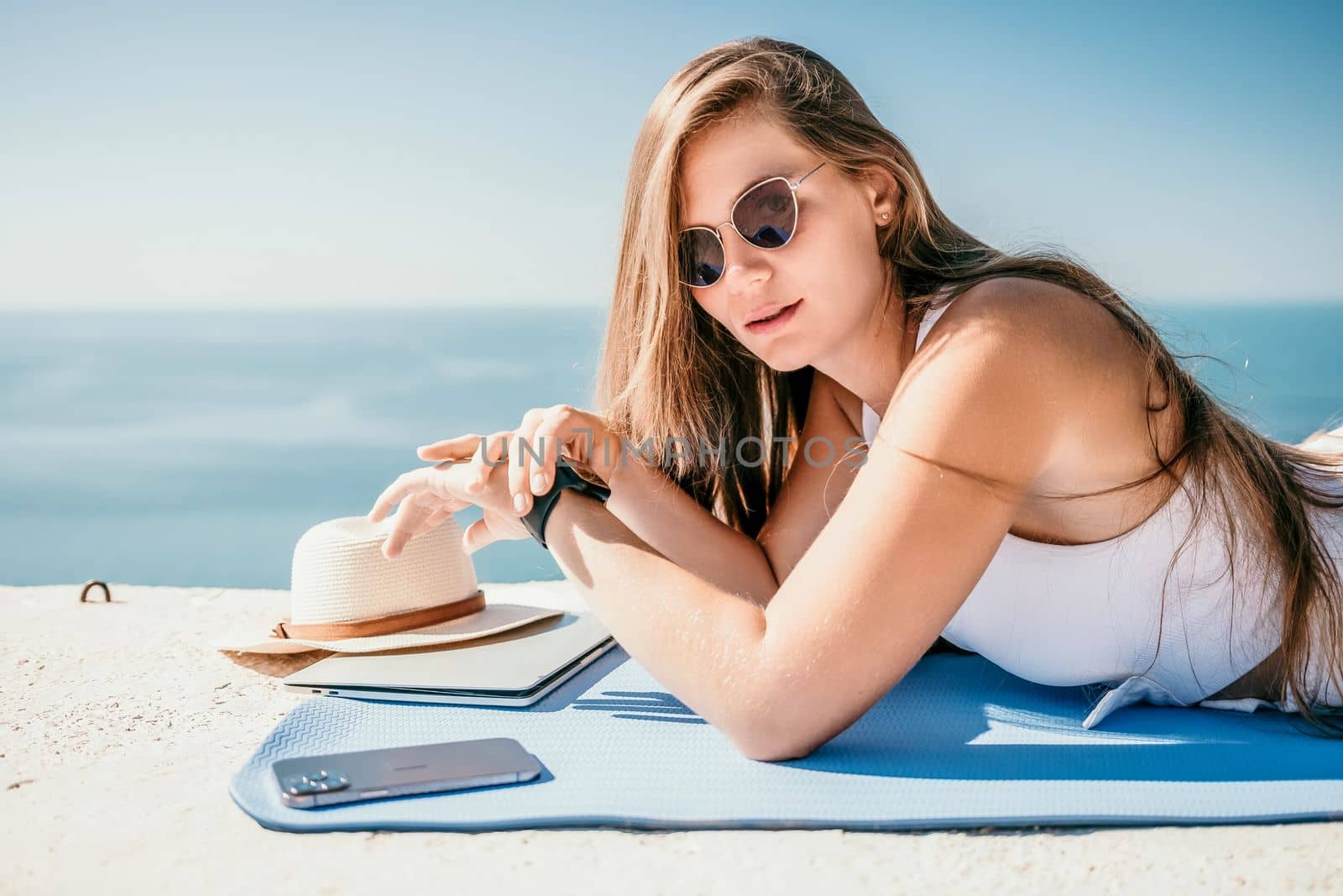 Successful business woman in yellow hat working on laptop by the sea. Pretty lady typing on computer at summer day outdoors. Freelance, travel and holidays concept.