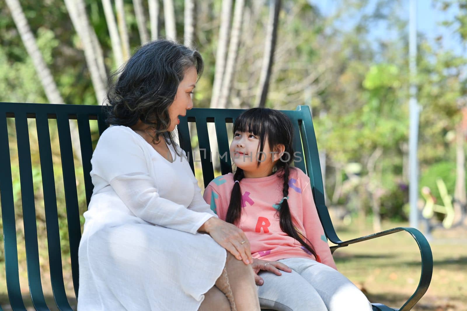 Happy little girl enjoy talking with elderly grandmother while sitting on a bench in the park. Family and love concept.
