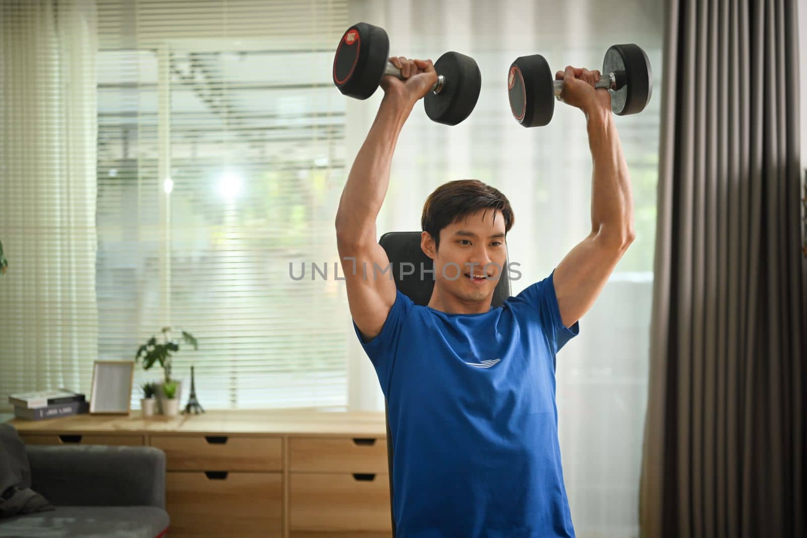 Strong athletic man doing dumbbell workout in bright living room. Fitness, sport and healthy lifestyle concept.