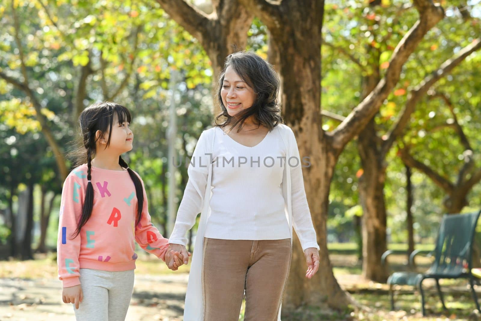 Smiling middle age grandmother and cute granddaughter walking in public park at morning by prathanchorruangsak