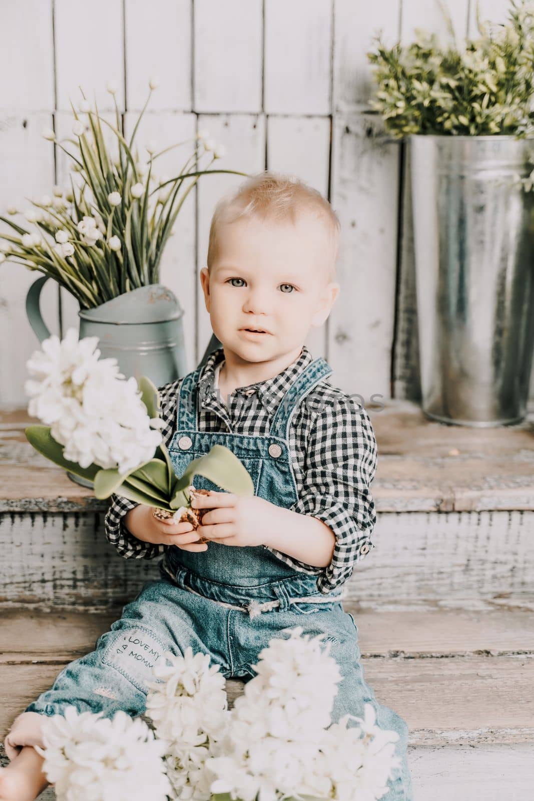 Girl 1 year old. sits holding a white hyacinth with a bulb in her hands, she is dressed in a plaid shirt and denim overalls on a wooden background with green plants in pots. by Matiunina