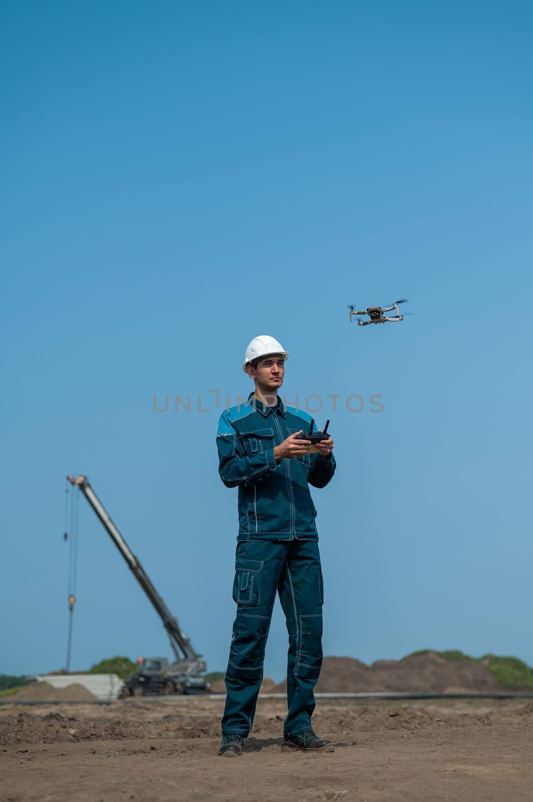 A man in a helmet and overalls controls a drone at a construction site. The builder carries out technical oversight