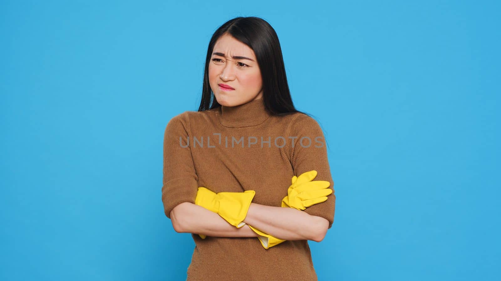 Nervous professional cleaner with protective yellow gloves having problem with house cleaning, standing with arm crossed in studio over blue background. Housekepper having negative expression