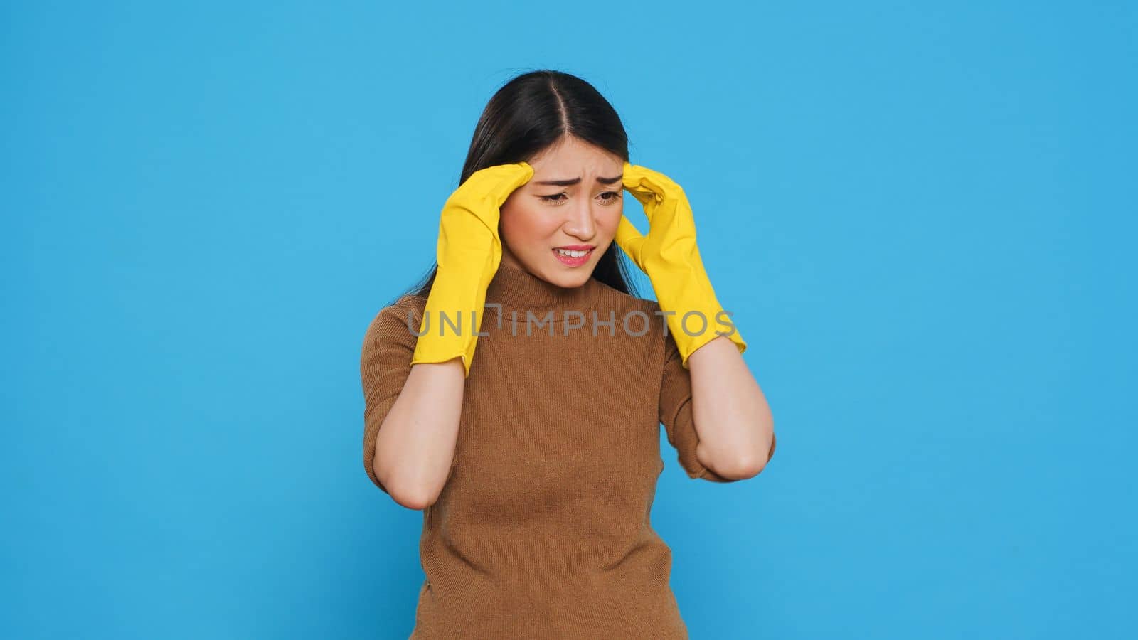 Overwhelmed housekeeper being tired after cleaning all house using variety of chemical detergents, standing in studio over blue background. Housewives relied on the cleaning lady services