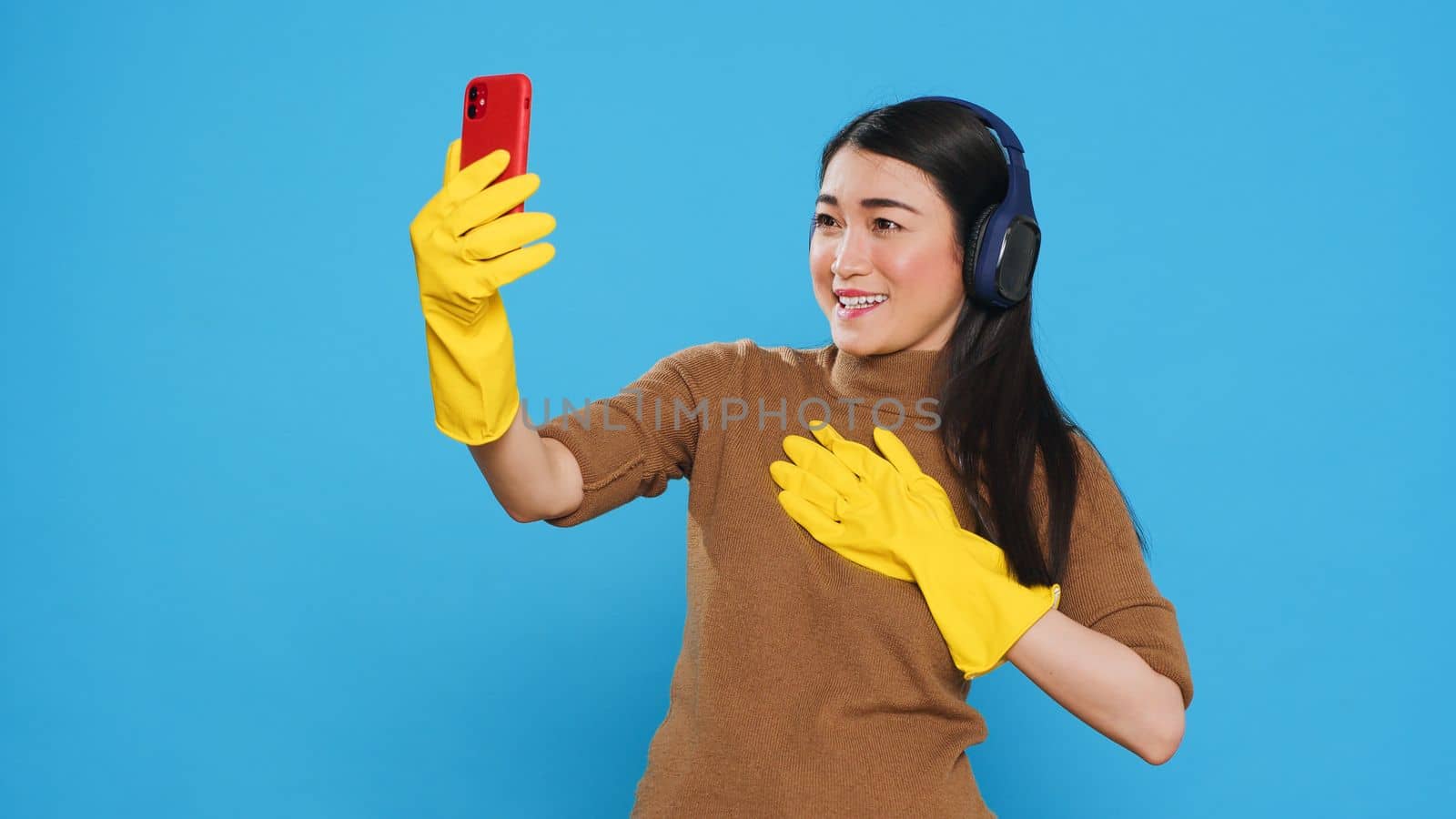 Smiling asian maid wearing headphone while talking with remote friend during videocall meeting on phone, standing in studio over blue background. Housekeeper doing house cleaning using chemical spray