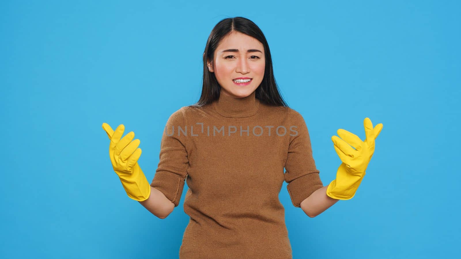 Nervous asian housekepper with protective gloves having problem with house cleaning, standing with arm crossed in studio over blue background. Maid having negative expression