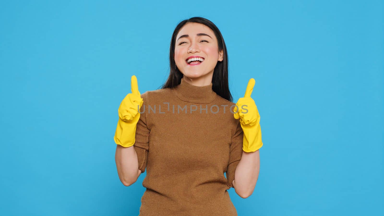 Positive joyful maid smiling while doing ok gesture after finishing to clean client house, posing in studio over blue background. Cheerful housekeeper is responsible for providing cleaning services