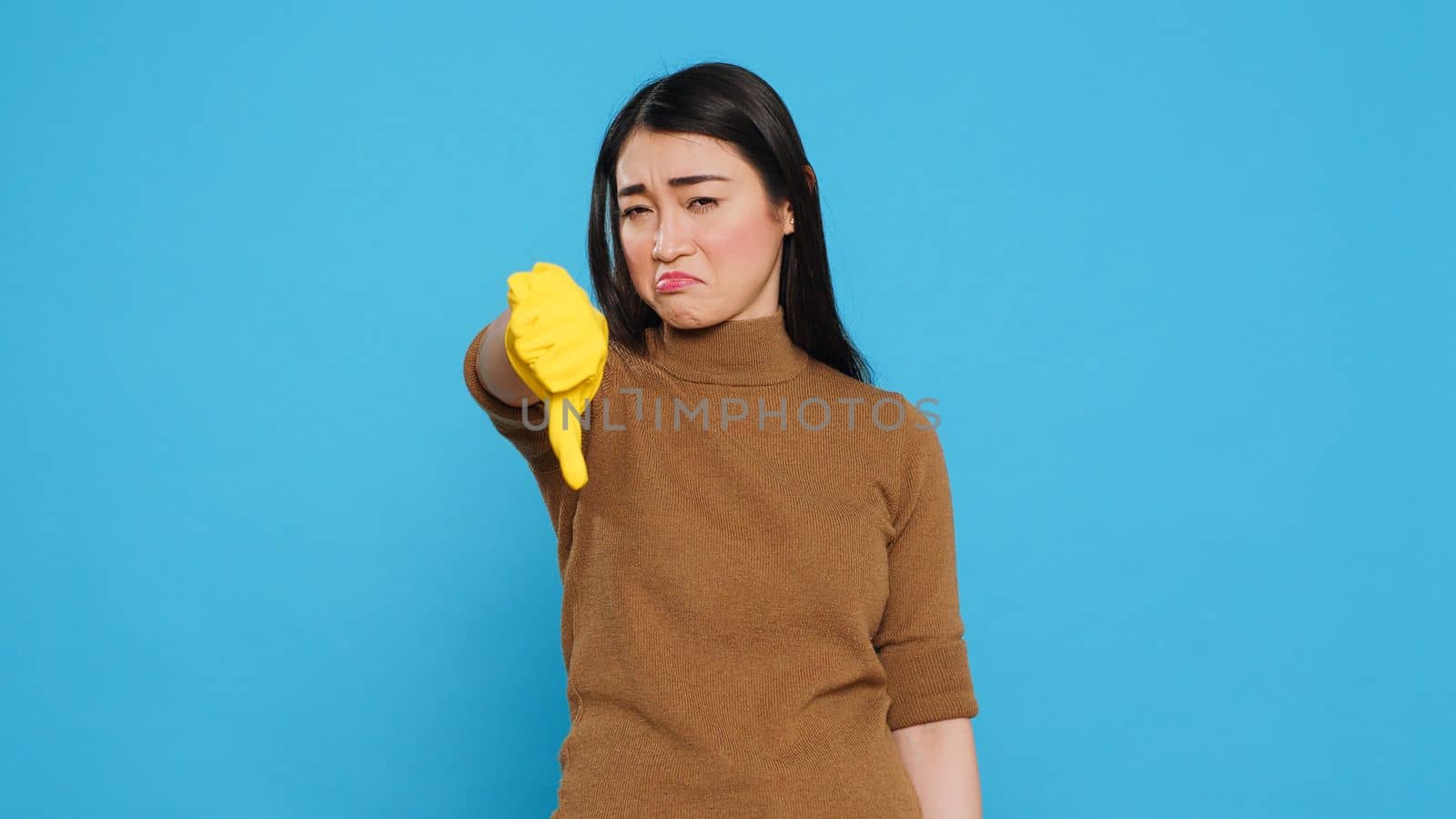 Upset asian maid doing thumbs down gesture while cleaning client house using houseclean detergent, posing in studio. Housekeeper is also knowledgeable about proper hygiene and cleanliness practices
