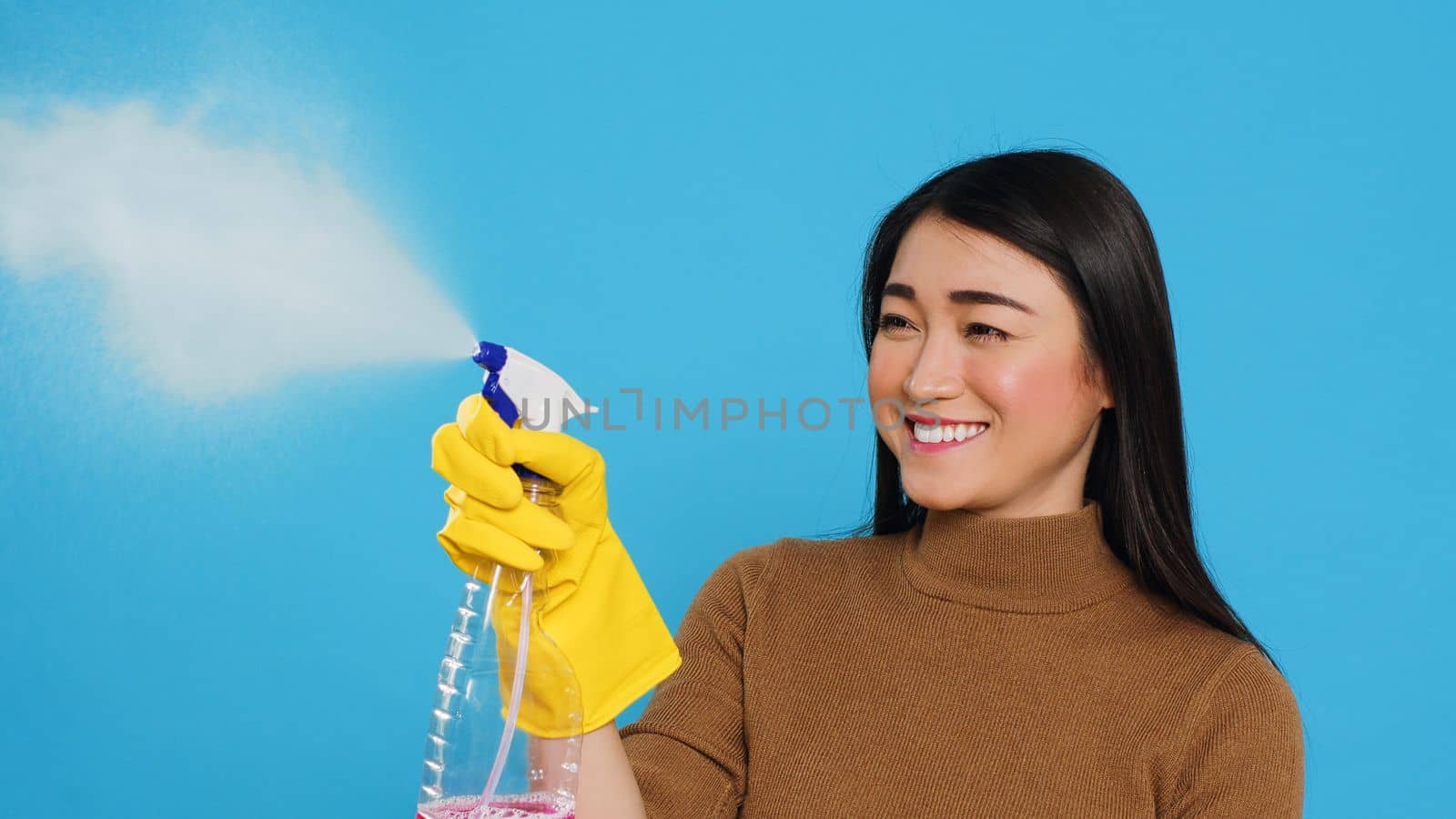 Housekeeper wearing yellow rubber gloves while holding spray and duster during houseclean, standing over blue background. Housewife primary responsibilities is to maintain cleanliness and hygiene