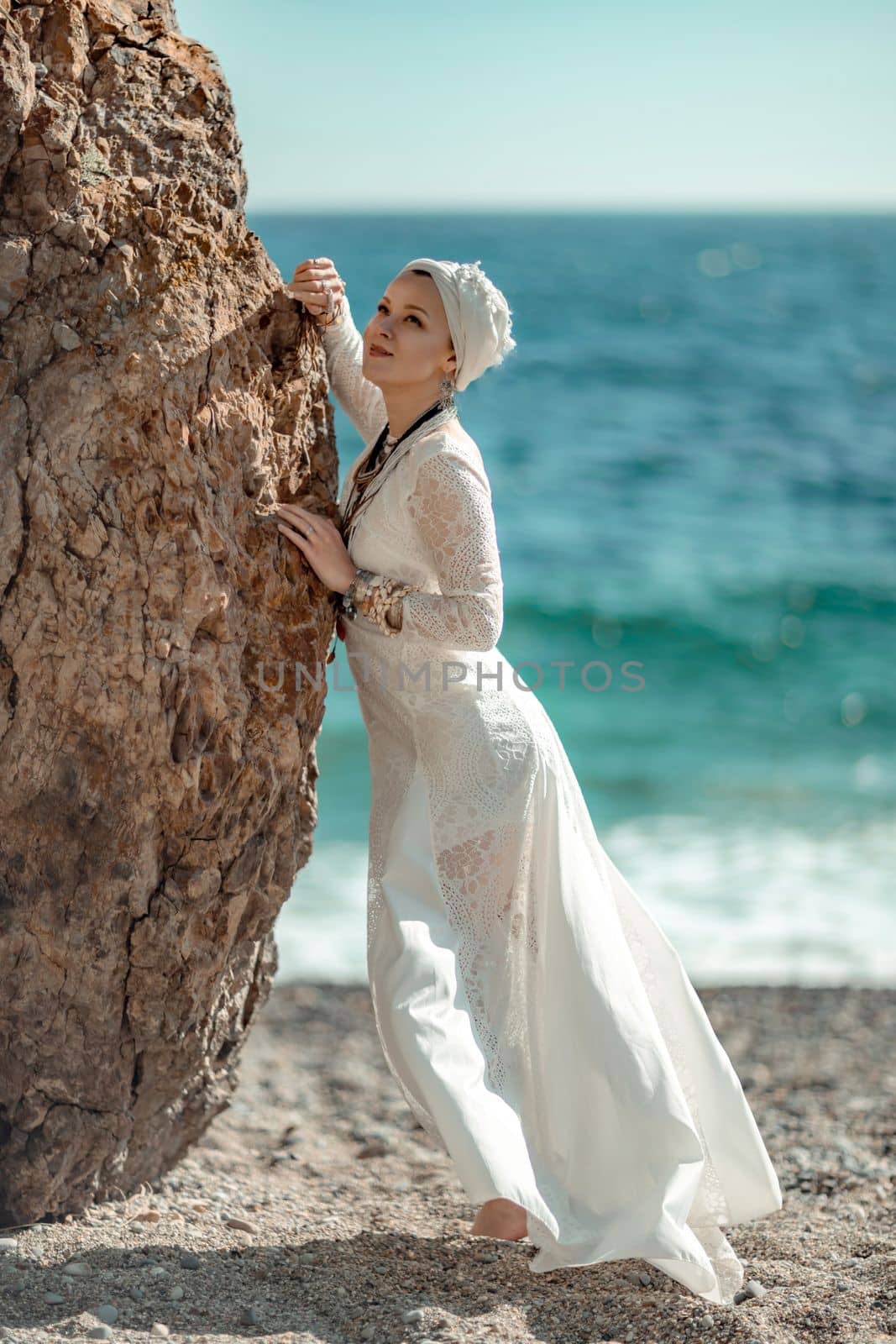 Woman white dress sea stones rocks.Middle-aged woman looks good with blond hair, boho style in a white long dress on beach jewelry around her neck and arms