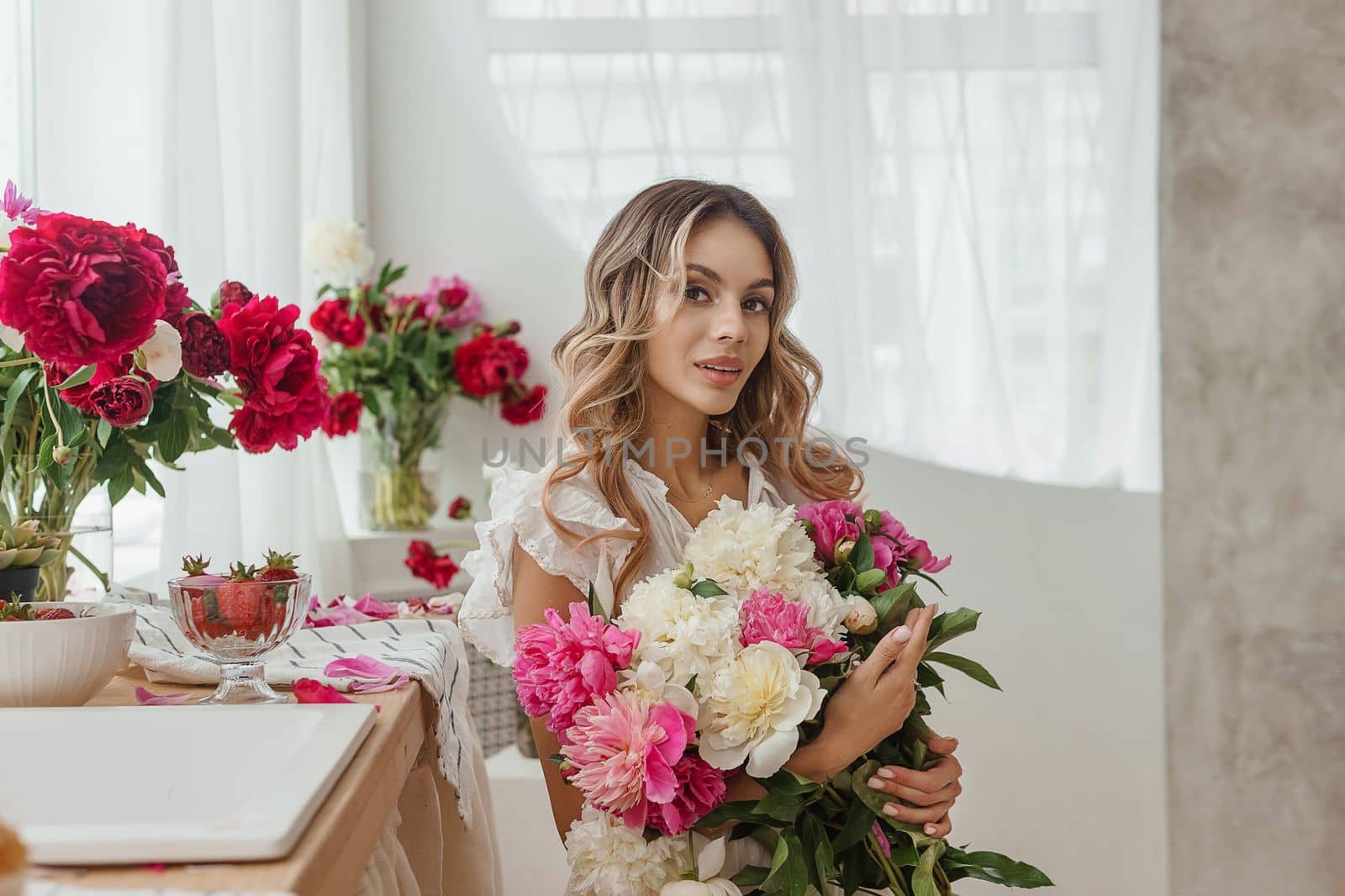 A beautiful girl in a white nightgown at home surrounded by spring flowers. A room decorated with bouquets of peonies