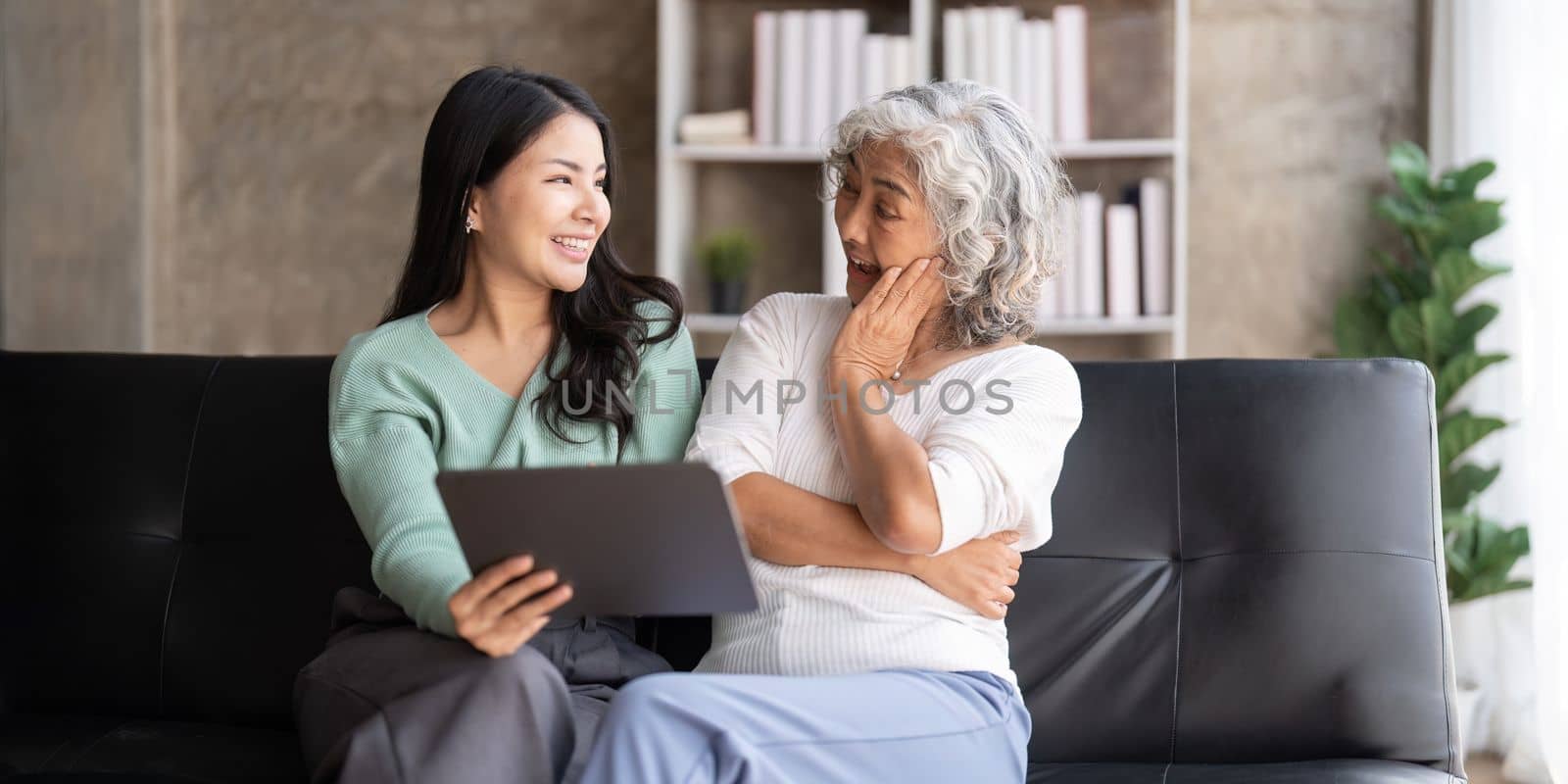 Mother up daughter sit on couch in living room hold on tablet looking at screen watching movie film, using device make video call, shopping ,daughter teach mom concept by nateemee