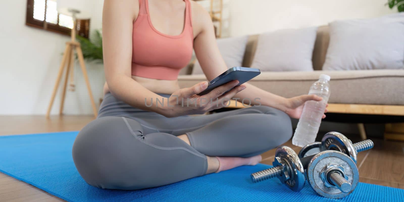 Woman sit on mat holding phone watching online workout training tv class video, using fitness app at home by nateemee