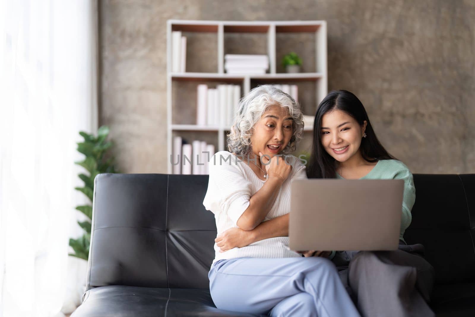 Mother up daughter sit on couch in living room hold on laptop looking at screen watching movie film, using device make video call, shopping website, daughter teach mom concept by nateemee