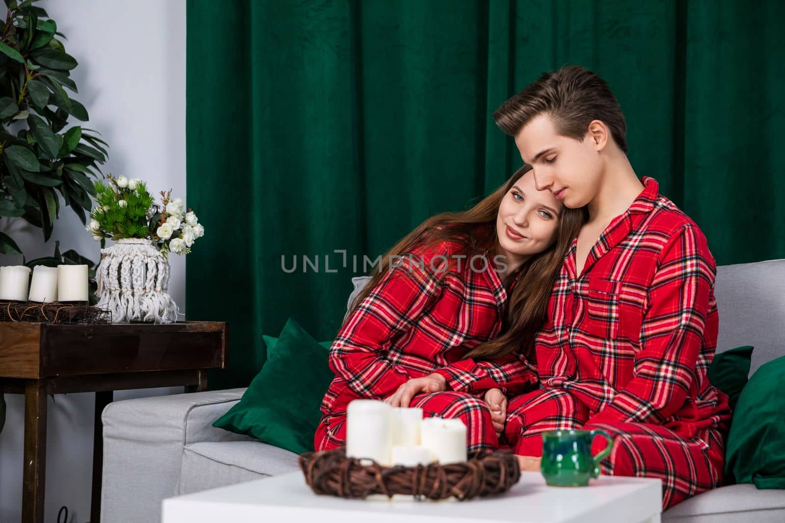 A couple in love posing for a session in a romantic interior. The wife cuddles up to her husband while sitting on a gray couch. In the background, behind the couch, is a heavy, dark green curtain