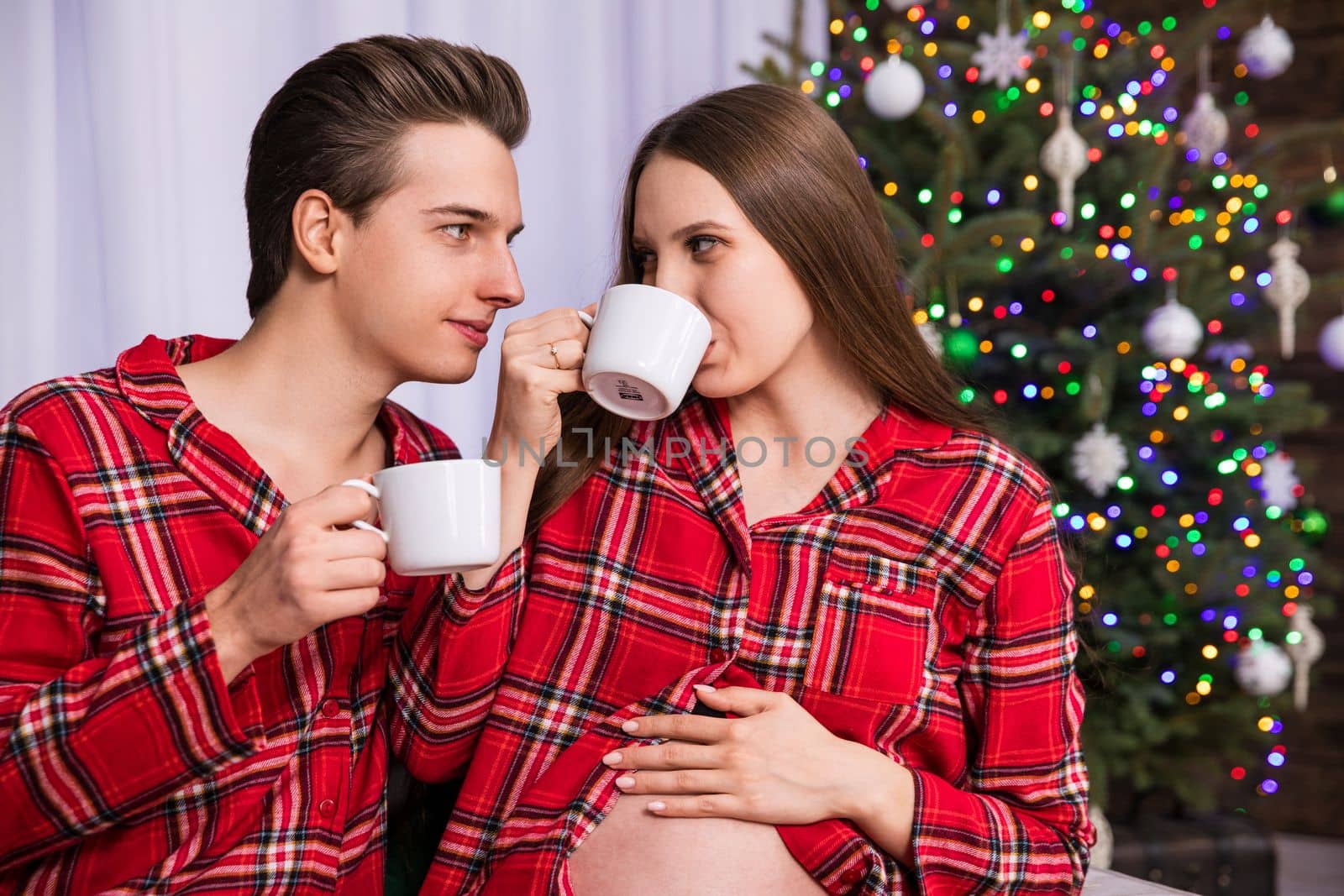 A man and a woman are sitting close together against the background of a fuzzy Christmas tree. The couple is holding white cups and the woman is drinking from a cup. The woman holds her hand over her exposed pregnant belly.