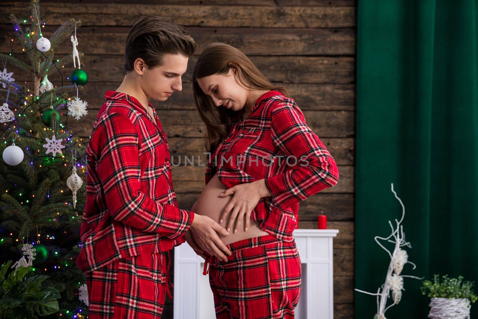 An expectant couple faces each other in a room with Christmas decorations and a Christmas tree. The man and the woman hold their hands on the pregnant belly.