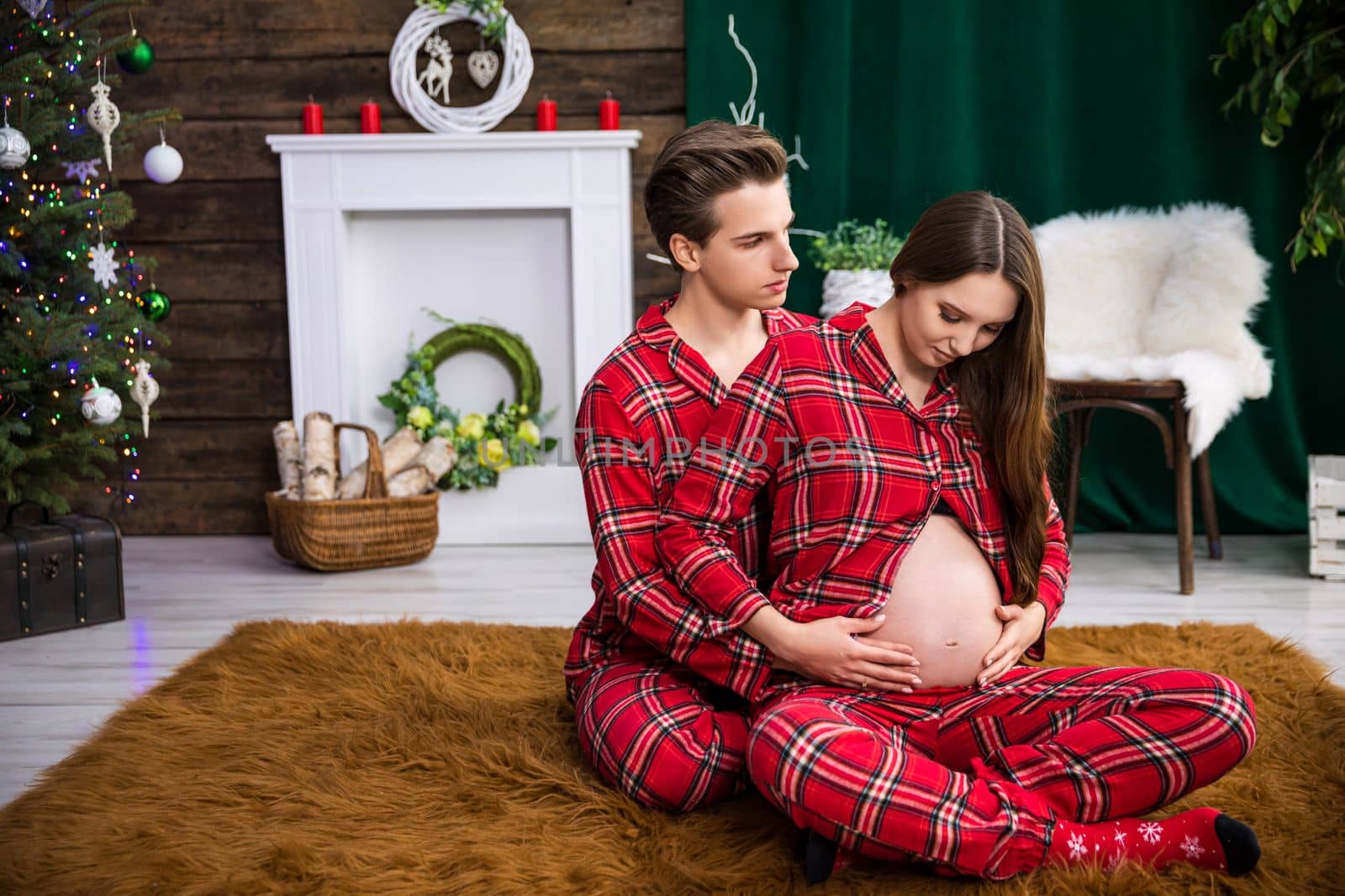 A man embraces a woman while sitting behind her back. The woman is advanced in pregnancy and exposes her exposed belly. Behind the couple in the background are Christmas decorations and a Christmas tree.