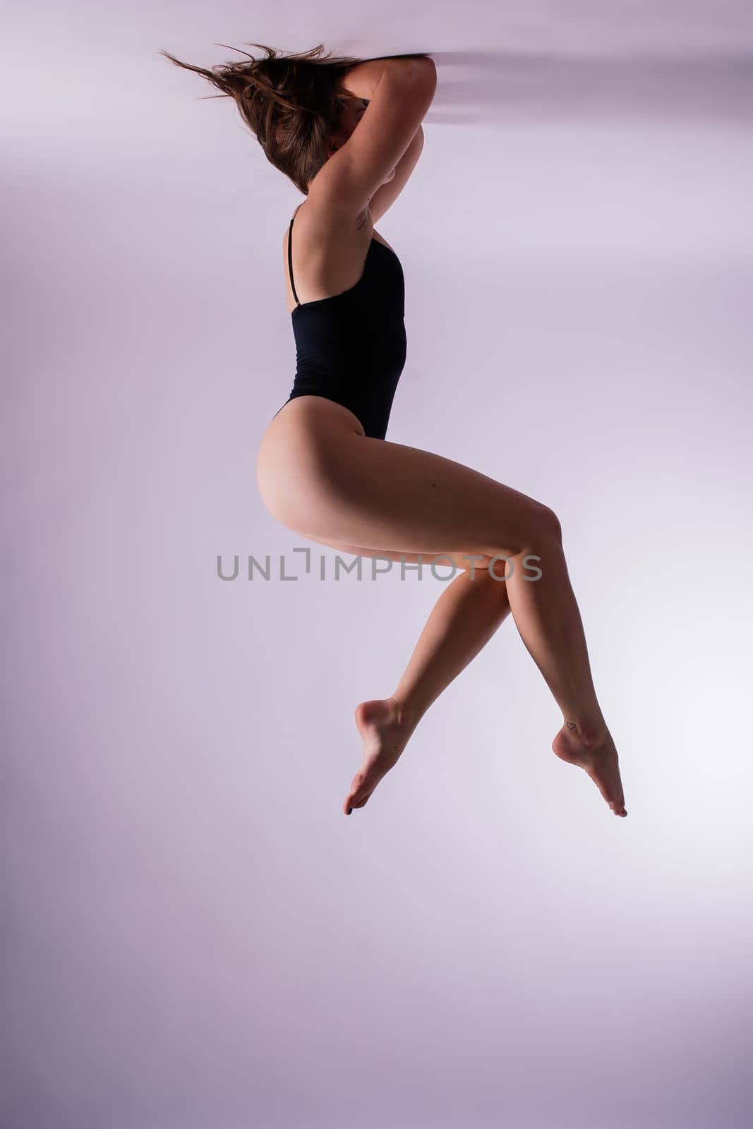 Young slim female in black bodysuit makes yoga stretching exercise on white studio wall background. by Zelenin