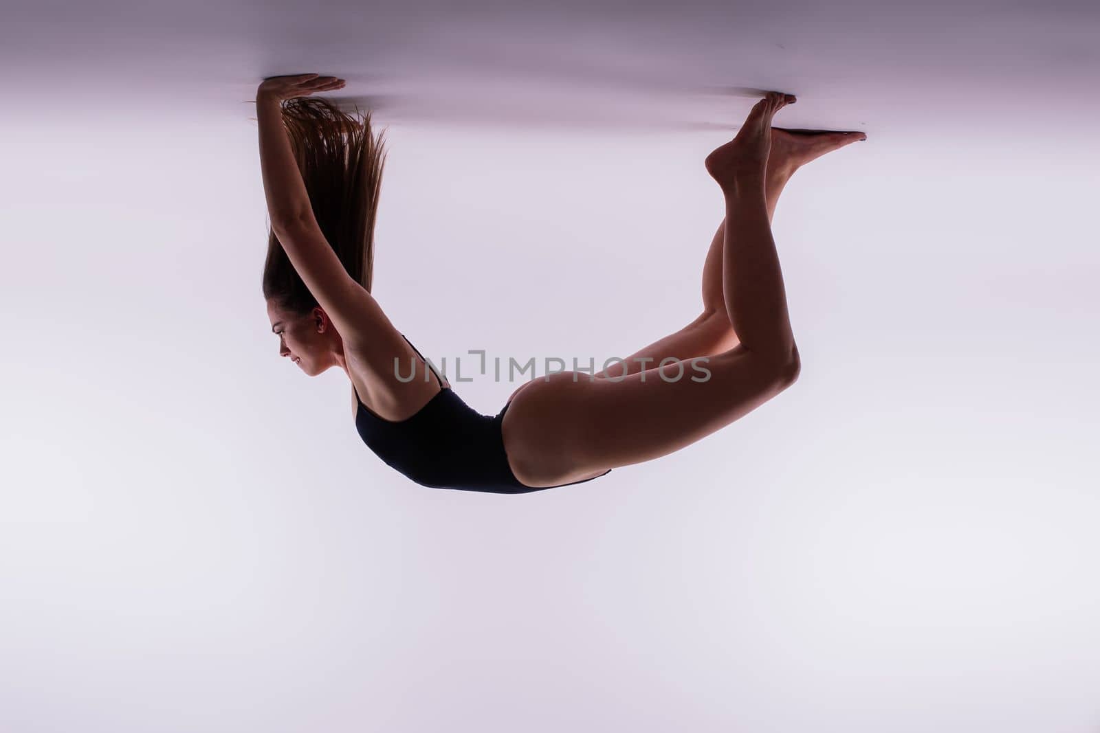 Young slim female in black bodysuit makes yoga stretching exercise on white studio background.