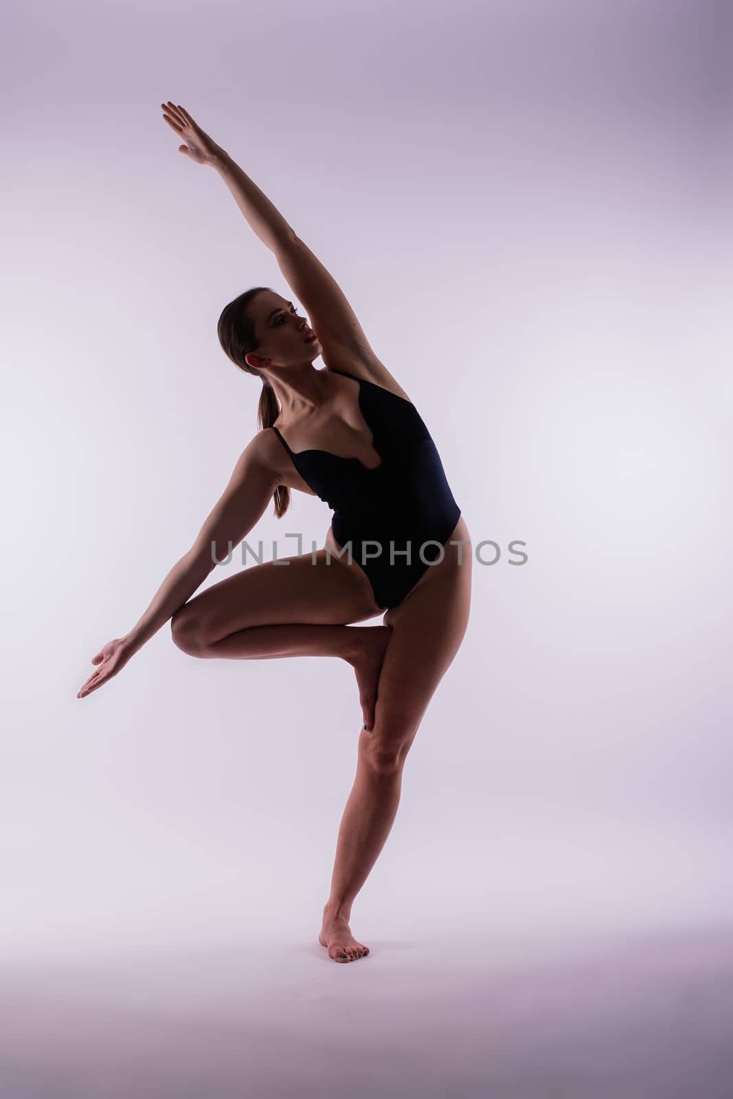 Young slim female in black bodysuit makes yoga stretching exercise on white studio wall background. by Zelenin