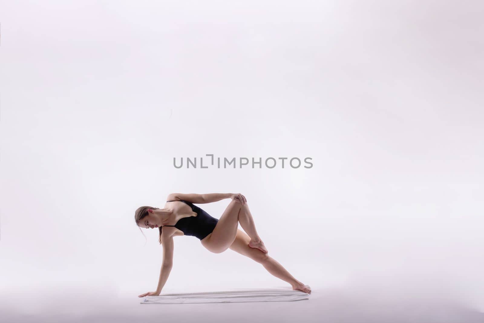 Young slim female in black bodysuit makes yoga stretching exercise on white studio background.