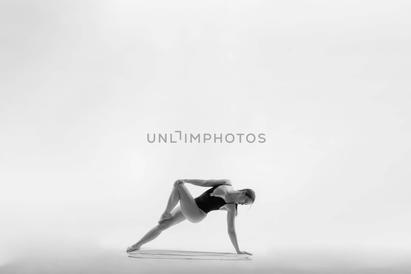 Young slim female in black bodysuit makes yoga stretching exercise on white studio background.