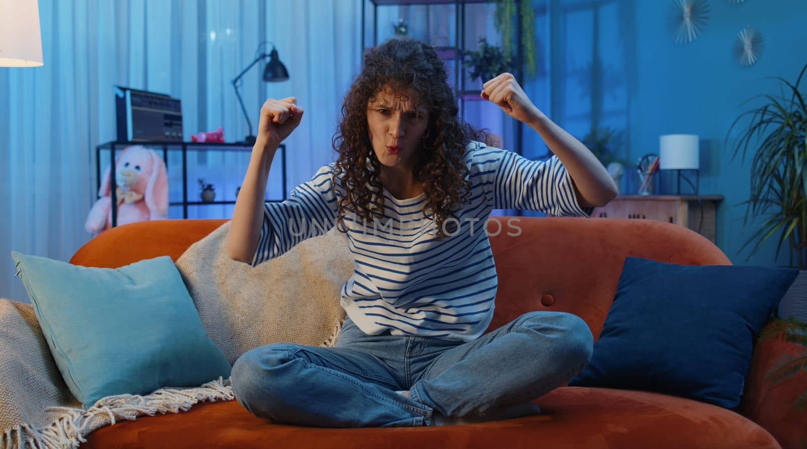 Happy woman with curly lush hairstyle shouting, celebrating success, winning, goal achievement, good news raising fists in gesture I did it, lottery luck at home apartment. Girl in night room on couch