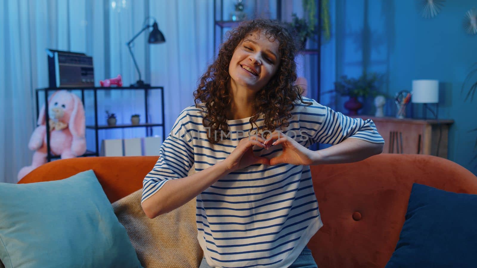 I love you. Happy woman with curly hairstyle at home couch makes symbol of love, showing heart sign to camera, express romantic feelings express sincere positive feelings. Charity, gratitude, donation