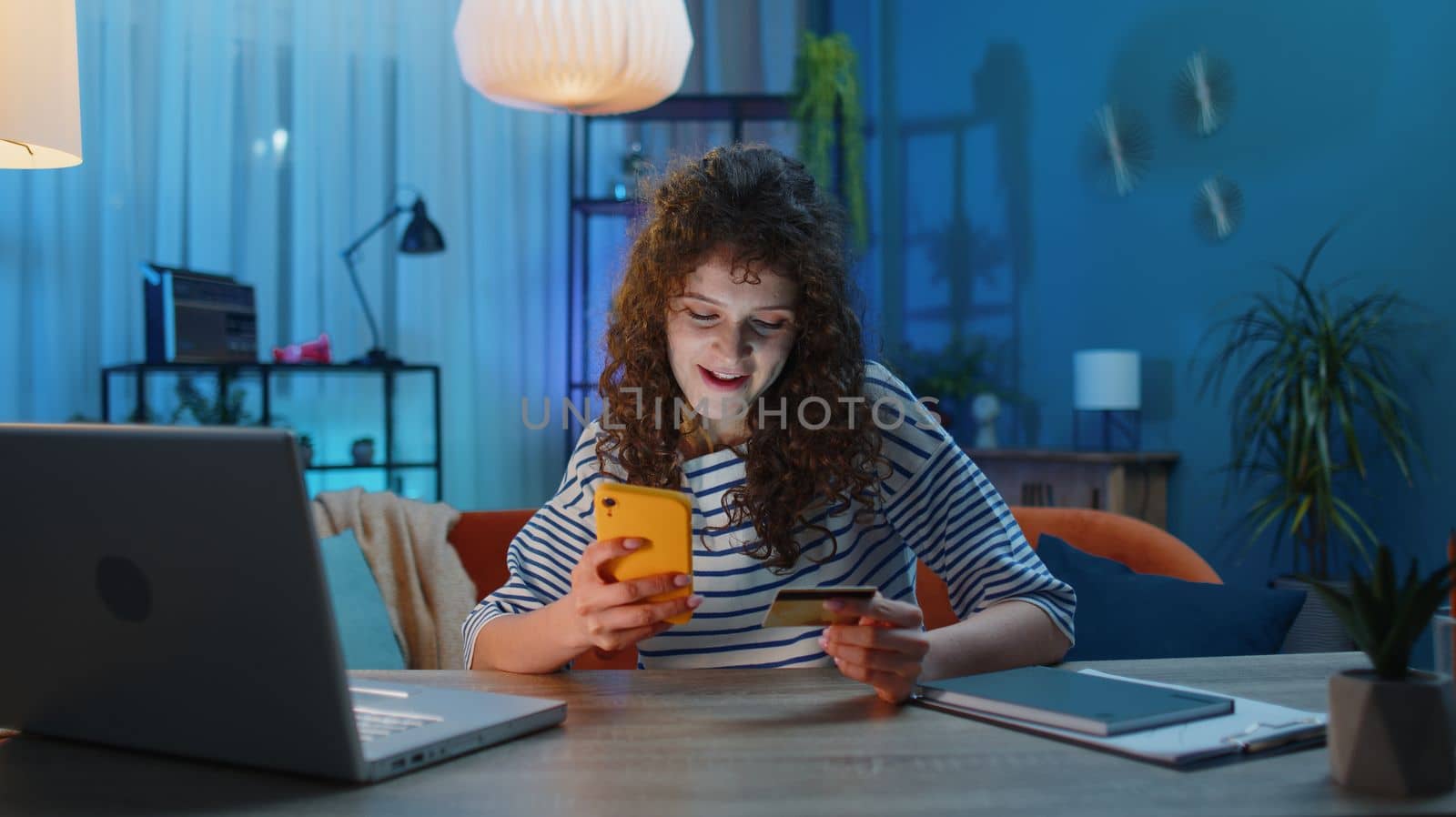 Young woman using credit bank card and smartphone while transferring money, purchases online shopping, order food delivery at home apartment indoors. Girl in evening room sitting at the office desk