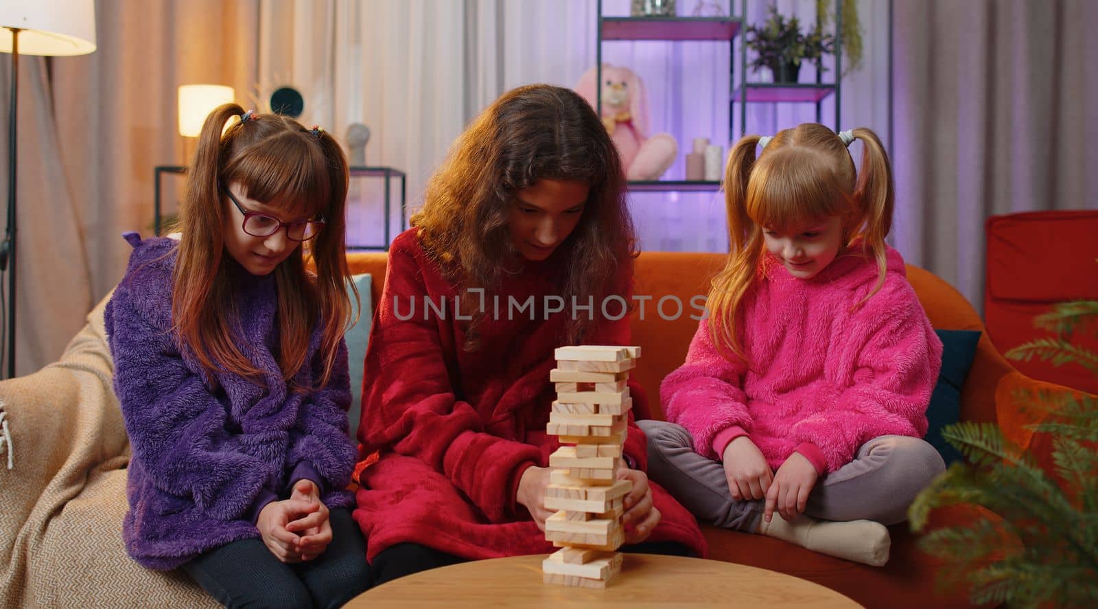 Teenage girl and toddler child sisters build tower from wooden bricks, losing board game competition. Three siblings children kids friends having fun with tower, playing with blocks at home playroom