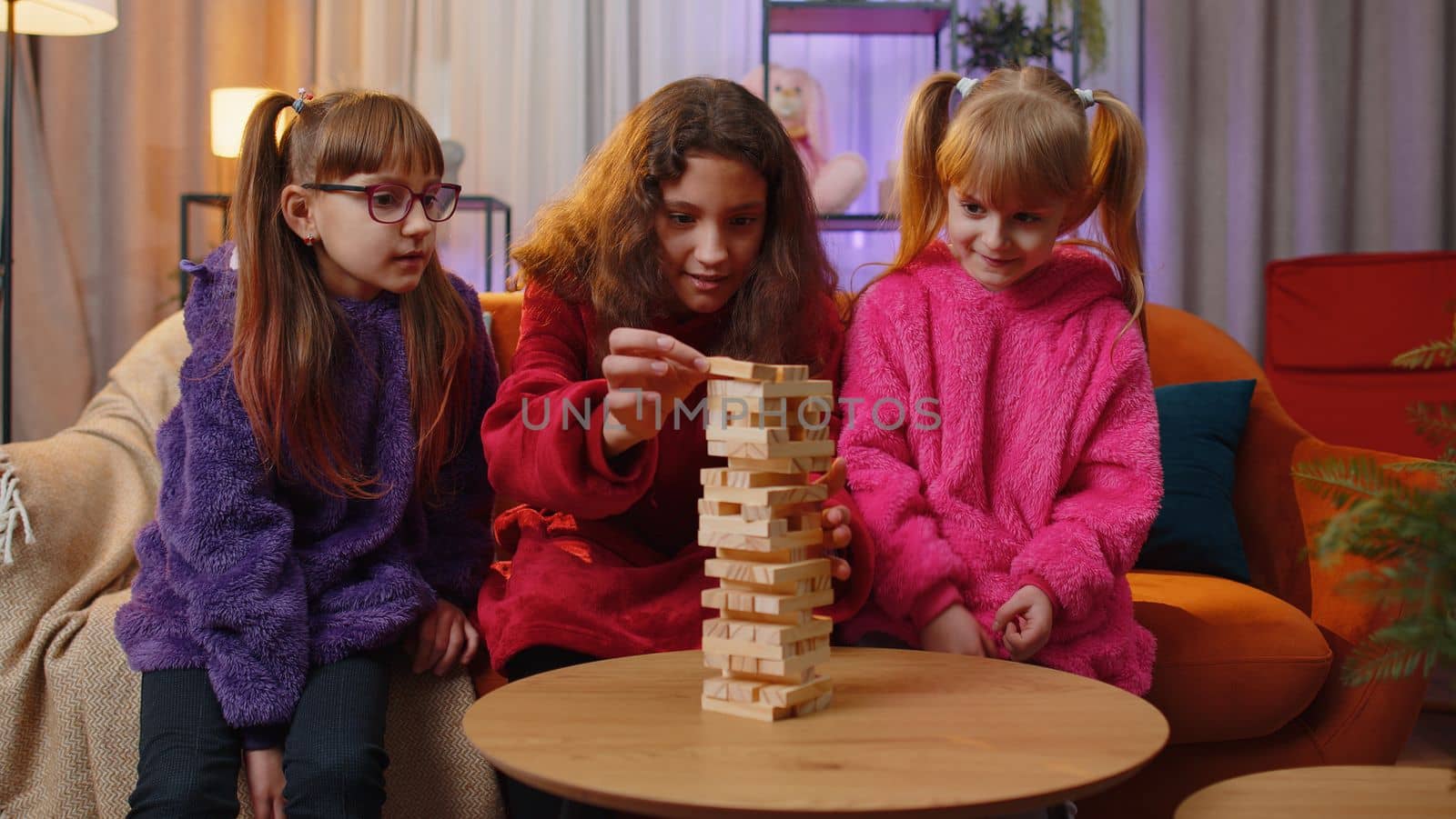 Teenage girl and toddler child sisters build tower from wooden bricks, losing board game competition. Three siblings children kids friends having fun with tower, playing with blocks at home playroom