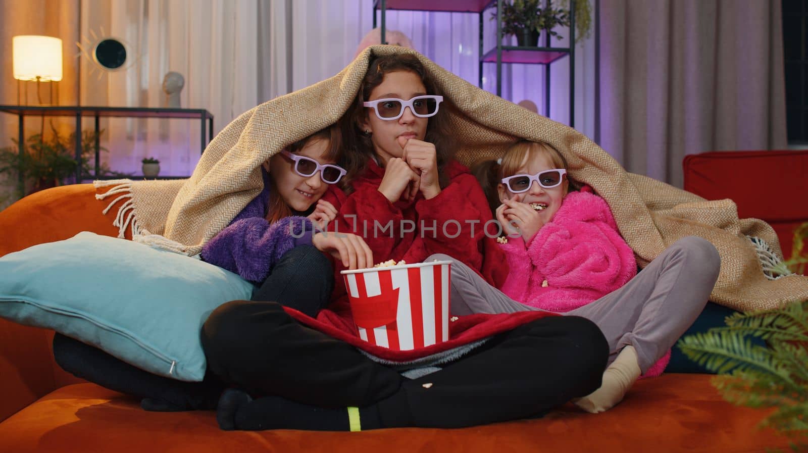 Excited teenage girl and little toddler child sisters under plaid eating popcorn and watching interesting tv serial, game, film, online social media movie. Three siblings children kids friends at home