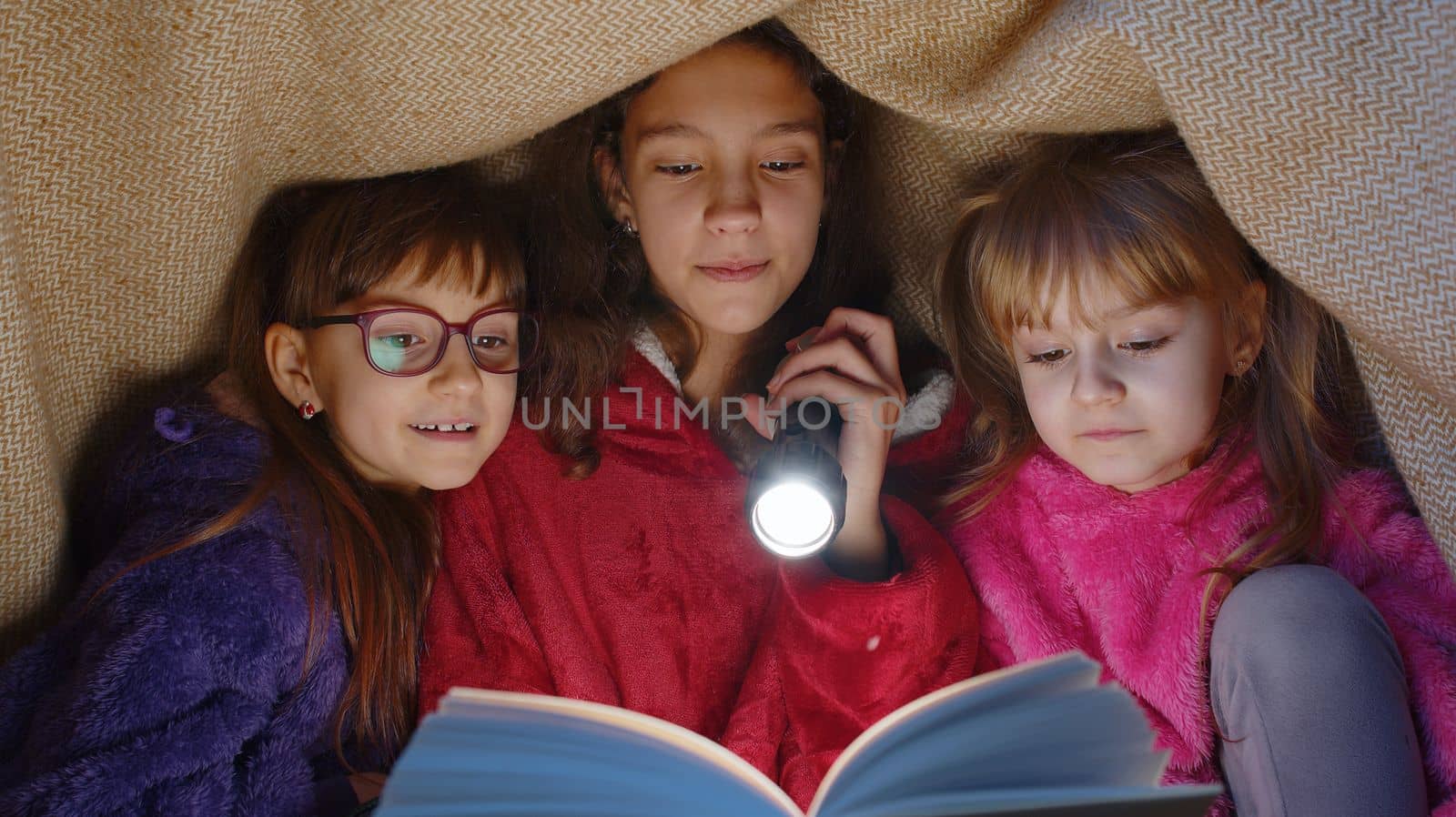 Teen girl and toddler child sisters under blanket plaid covers with flashlight reading interesting fairytale story book using flashlight. Female three siblings children kids friends in wigwam at home