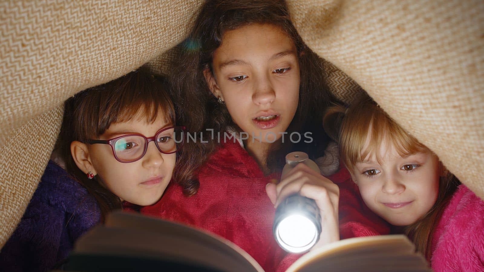 Teen girl and toddler child sisters under blanket plaid covers with flashlight reading interesting fairytale story book using flashlight. Female three siblings children kids friends in wigwam at home