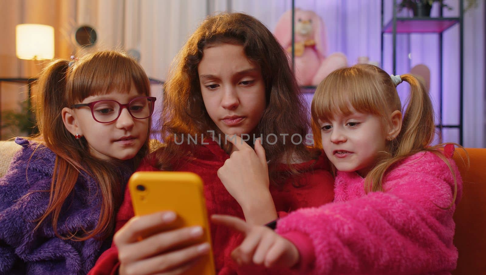 Teen girl, toddler child sisters using mobile phone texting share messages content on smartphone social media applications online, watching relax movie. Three siblings children kids at home playroom