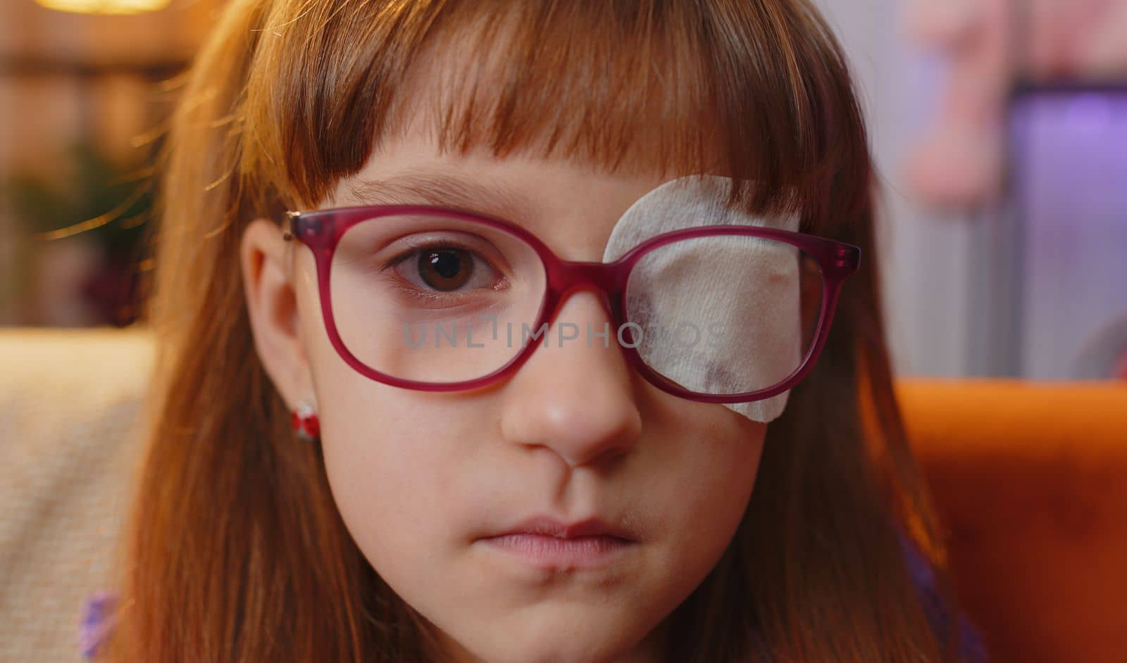 Sick ill toddler girl face looking at camera having protective bandage plaster patch scratch on one eye. Injury trauma blind. Child kid wearing glasses at home in room. Medical eyesight vision therapy