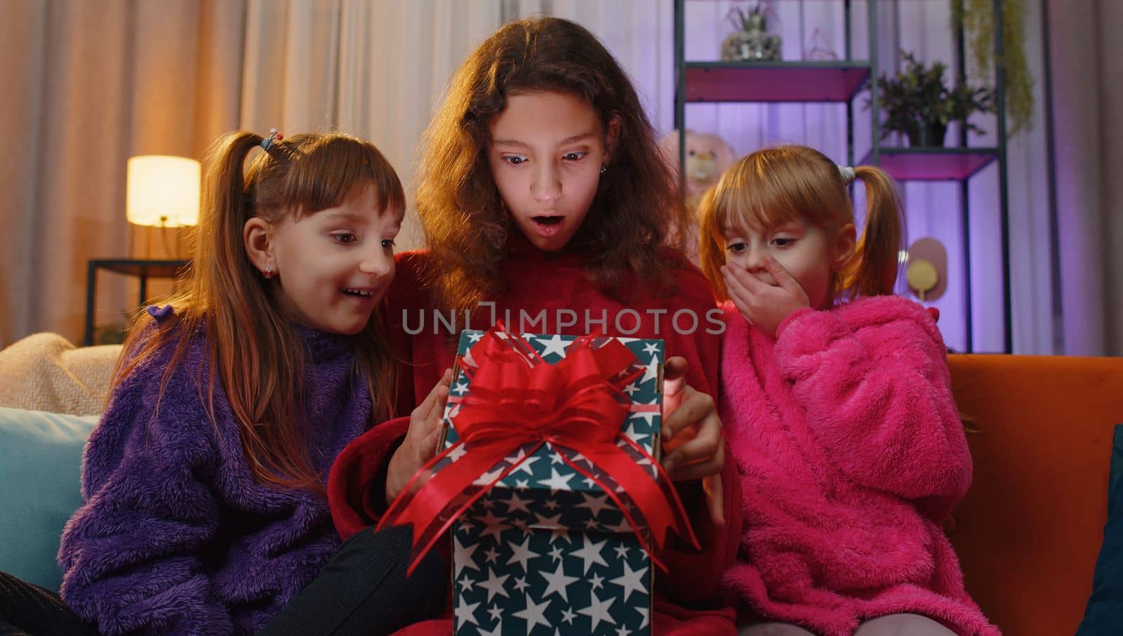 Happy teen girl and toddler child sisters opening Christmas gift box with excited surprised face. Female three children kids siblings or friends holding light glowing birthday present at home playroom