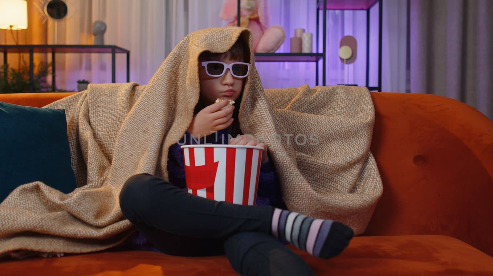 Excited toddler girl in 3D glasses eating popcorn and watching interesting tv serial, sport game, film, online social media movie content. Teen child kid at home in evening living play room on couch