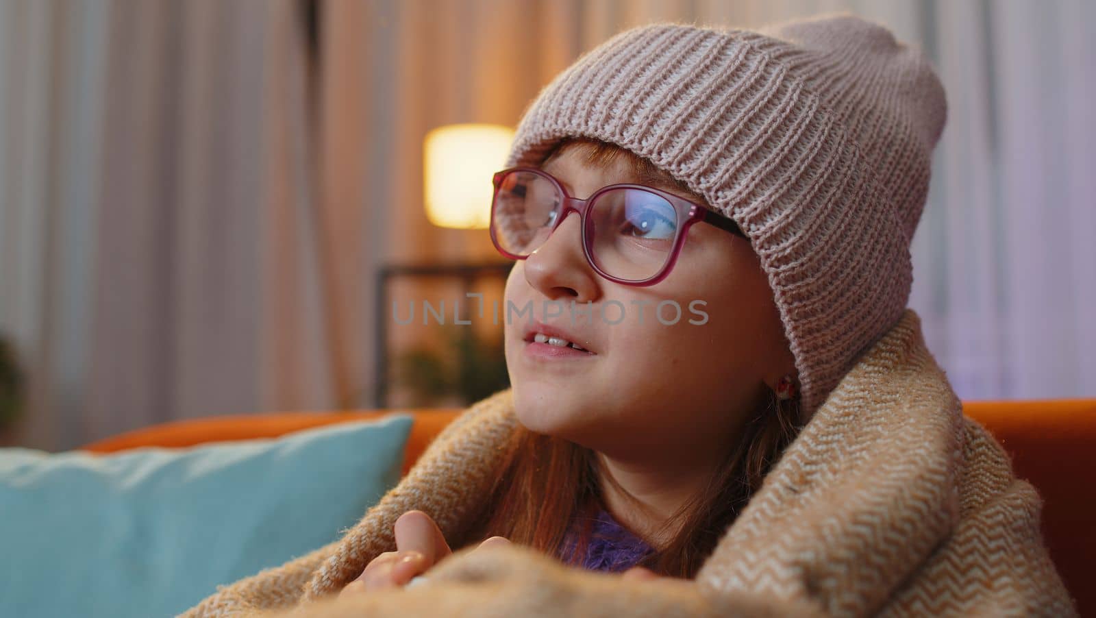 Sick toddler girl wear hat wrapped in plaid sit alone shivering from cold on sofa drinking hot cacao in unheated apartment without heating due debt. Unhealthy kid feeling discomfort try to warming up