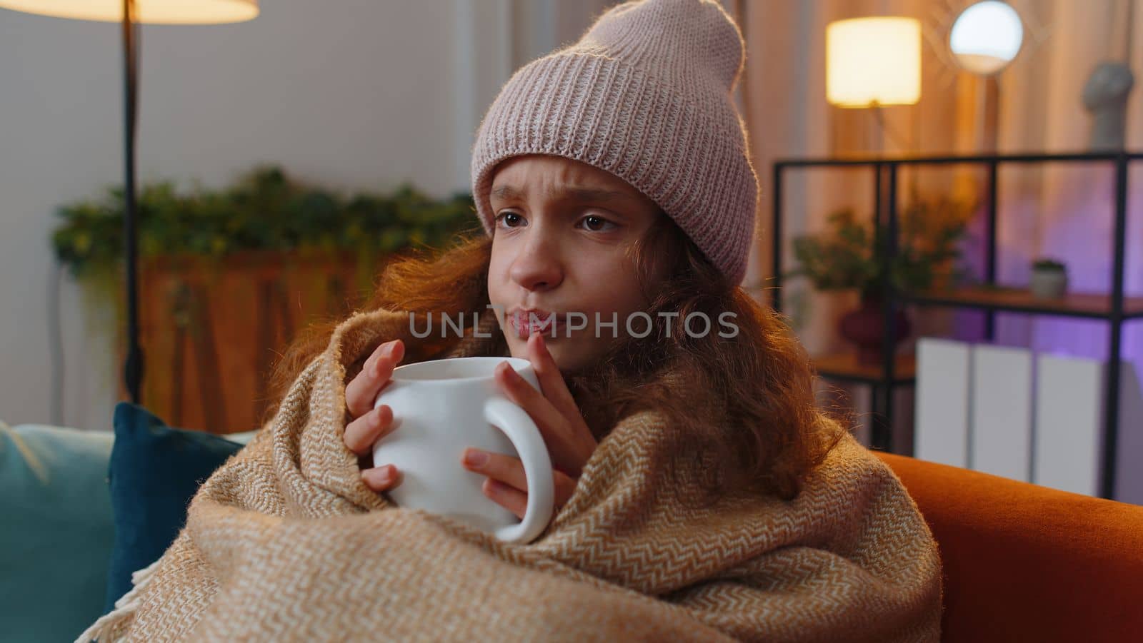 Sick teenager girl wear hat wrapped in plaid sit alone shivering from cold on sofa drinking hot cacao in unheated apartment without heating due debt. Unhealthy kid feeling discomfort try to warming up